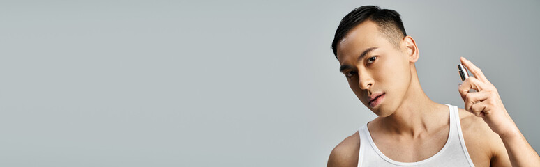 A handsome Asian man in a tank top spraying perfume in a grey studio.