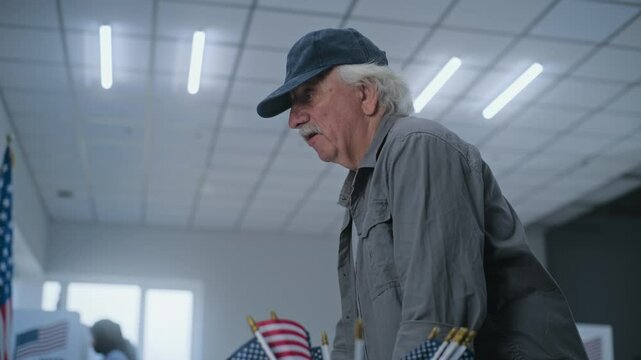 Caucasian elderly US citizen stands near registration table at polling station and talks to polling officer. Female worker consults man and gives ballot paper for voting. US presidential elections.