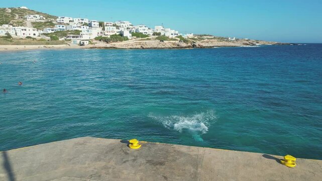 A boy stands on a pier at Stavros Beach in Donousa, Greece, ready to jump into the crystal-clear waters. Village houses and a sandy beach are visible in the background