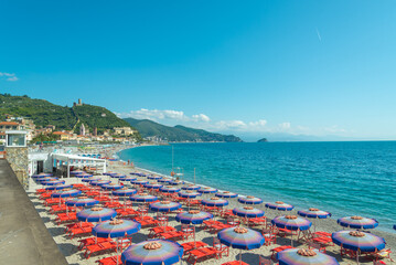 The coastline of Noli, Liguria, Italy, with beach umbrellas in several rows on the sand. Turquoise waters of mediterranean sea on the right. Green hills on the background.