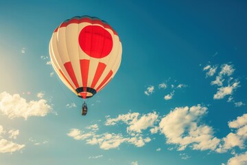 Fototapeta premium A hot air balloon adorned with the Japanese flag ascends against a backdrop of fluffy white clouds and a bright blue sky.