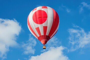 Fototapeta premium A red and white hot air balloon, featuring the Japanese flag design, rises gracefully against a backdrop of bright blue sky and puffy white clouds.