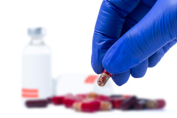 Doctor's hand holding red pill in blue gloves in front of vaccine medical vials and pills with focus outs on white background. Closeup