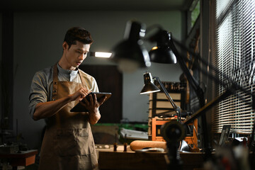 Professional artisan wearing an apron using a digital tablet in his workshop. Small business and technology concept