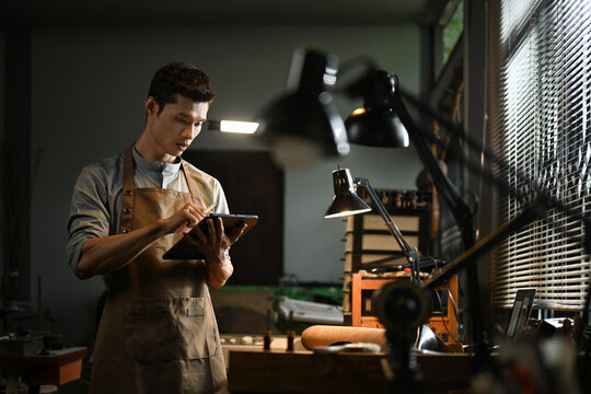 Professional artisan wearing an apron using a digital tablet in his workshop. Small business and technology concept - Powered by Adobe