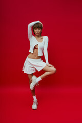 A stylish young woman in a short skirt and crop top energetically dancing on a vibrant red backdrop.