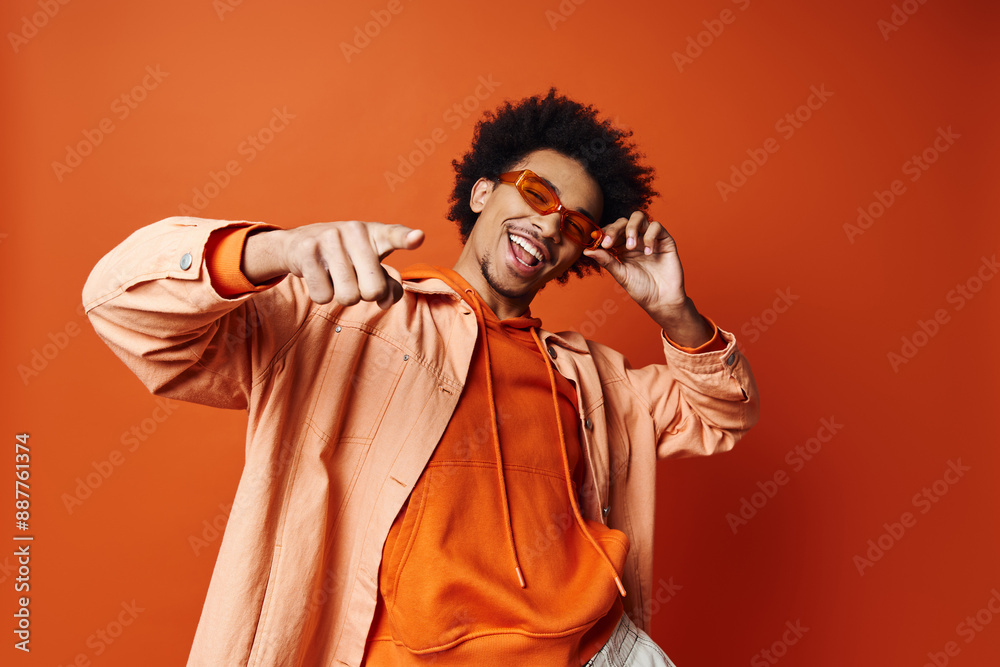 Wall mural a stylish young african american man with curly hair wearing an orange shirt and sunglasses, posing 