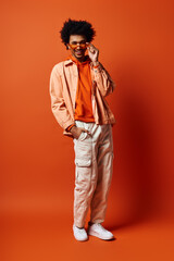 A trendy, emotional young African American man stands confidently against a bright orange background in fashionable attire and sunglasses.