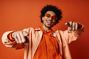 Stylish African American man in orange shirt and sunglasses pointing towards camera on vibrant...