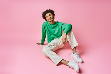 A cheerful African American man with curly hair sitting on a pink surface, wearing a green shirt...