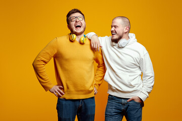 Obraz premium Two excited friends in casual wear standing and laughing together. Best friends enjoying isolated over yellow background. Two men with headphones having fun in studio. Friendship concept