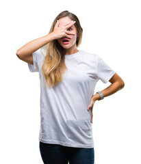 Young beautiful blonde woman wearing casual white t-shirt over isolated background peeking in shock covering face and eyes with hand, looking through fingers with embarrassed expression.