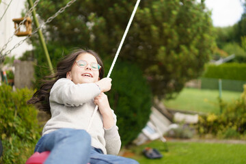 A girl with glasses with curly hair rides on a swing in the backyard in the summer. Happy childhood concept.