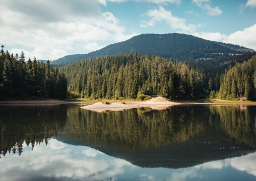Synevyr lake in Carpathian Mountains. Lake is surrounded by high pine trees. Reflection of the forest and hill range in the water. Partly cloudy sky in the background.