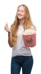 Blonde teenager woman eating pop corn with a happy face standing and smiling with a confident smile showing teeth