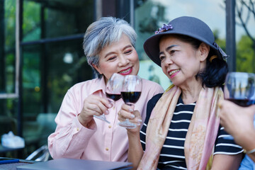 Elderly Asian women sitting at a restaurant's outdoor seating, raising glasses of red wine. Casual attire, cheerful expressions, lush greenery in the background, celebrating friendship