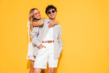 Happy beautiful romantic teenage couple. Smiling handsome caucasian man and young woman in summer clothes posing in studio, hugging sharing emotions, cheerful and happy. Isolated on yellow, sunglasses