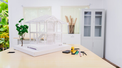 Desk of an engineer displays a concrete house structure under construction. A toy house model sits nearby, surrounded by blueprints and tools, depicting the foundation, roof, and structural elements.