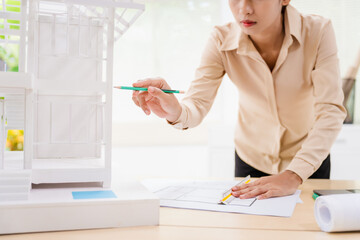 An Asian architect woman sits at her desk, wearing a yellow safety hat. A concrete house structure...