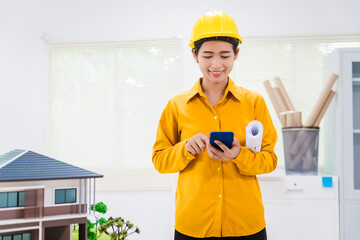 An Asian architect woman sits at her desk, wearing a yellow safety hat. A concrete house structure and toy model depict a hip roof house with various rooms and a garden design.