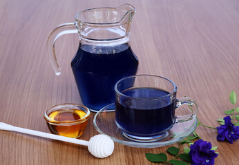 Purple water in glass cups and pitchers Honey with a scoop and purple clitoria ternatea flowers all on a large wooden table.

