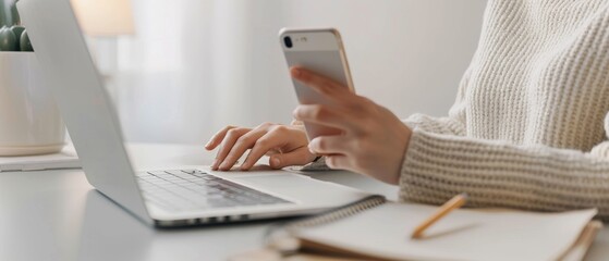 Fototapeta premium A person sitting at their desk, holding an iPhone in one hand and using the other to type on a laptop keyboard.
