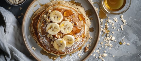 A top view photograph featuring a breakfast spread of fluffy pancakes topped with sliced bananas drizzled honey and sprinkled oats on a plate placed on a table with a napkin offering ample copy space