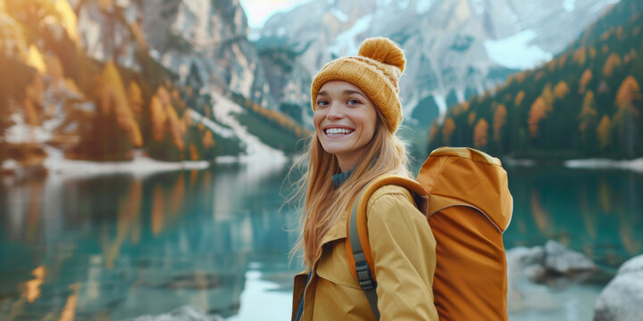 Cheerful young woman in beanie and backpack enjoys hike by serene mountain lake surrounded by autumn foliage
