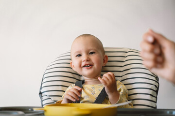 A baby girl sits in a high chair and is being fed by mother. The baby is wearing a yellow and white floral pattern shirt and has her hands on the tray of the high chair.