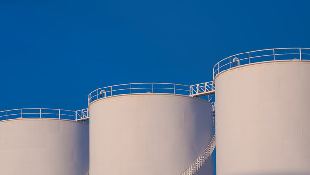Row of 3 white storage fuel tanks against blue clear sky background, low angle view with copy space