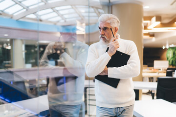 elderly Caucasian business man with white hair and beard stands thoughtfully in modern office....