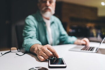 Elderly 60 years old Caucasian man using banking app on his smartphone at desk with a laptop,...