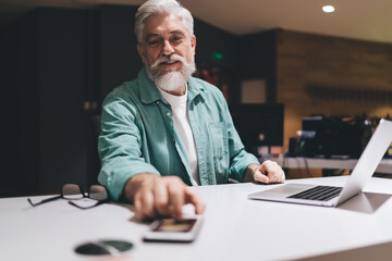 Elderly Caucasian man with white hair and beard, using smartphone banking application for financial payment on desk with laptop, glasses. Professional office setting, evening time, focused expression