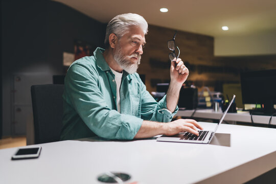 Elderly Caucasian man with gray hair and beard typing on laptop in office. Businessman holding his glasses, deep in thought. analyzing financial reports, working on creative project late into evening - Powered by Adobe