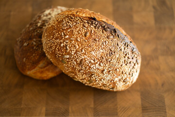 Two loaves of sourdough bread, freshly baked and still warm, are stacked on a wooden surface. The loaves are covered in seeds sesame and sunflower, adding a rustic and flavorful touch.