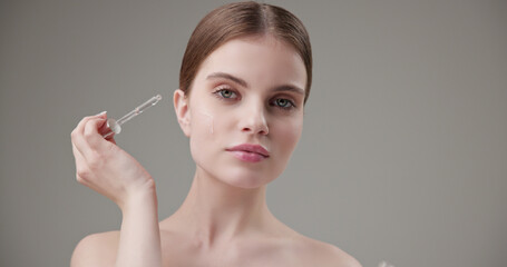 Fototapeta premium Beauty, health, cosmetics, anti-aging therapy and skin care concept - young beautiful brunette Caucasian woman applying cosmetic serum from a pipette for facial skin care