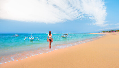 Obraz premium A young girl in a bikini walking on the beach - Tropical sandy beach and clear sea background with traditional balinese boats - Bali, Indonesia 