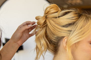 Fototapeta premium Professional stylist styles the hair of blonde woman. Styling hairstyles for wedding or event. High bun of curly hair