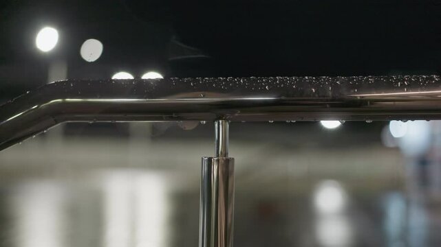 Forward view of a rain-drenched metal handrail at night, with water droplets glistening under soft lighting