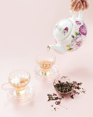 Fancy tea pot pouring rose tea into glass tea cups on pink background 