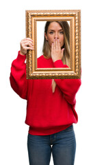 Fototapeta premium Beautiful young woman holding vintage frame cover mouth with hand shocked with shame for mistake, expression of fear, scared in silence, secret concept