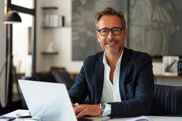 A professional businessman seated at a desk with a laptop, smiling confidently, surrounded by modern office decor and a large abstract painting in the background.