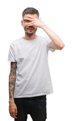 Fototapeta premium Young tattooed adult man peeking in shock covering face and eyes with hand, looking through fingers with embarrassed expression.