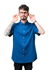Fototapeta premium Young handsome man wearing glasses over isolated background Smiling pulling ears with fingers, funny gesture. Audition problem