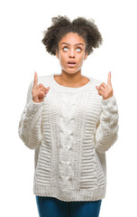 Young afro american woman wearing winter sweater over isolated background amazed and surprised looking up and pointing with fingers and raised arms.
