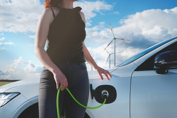 Side view on white electric car with plugged in green charging cable at wind park. Woman with painted nails holds cable while waiting. Focus on green glowing charge port. Modern transportation concept