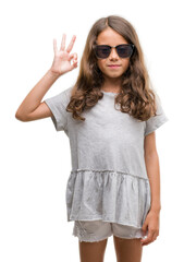 Naklejka premium Brunette hispanic girl wearing sunglasses smiling positive doing ok sign with hand and fingers. Successful expression.