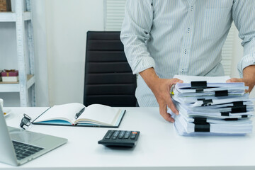 employee manages a stack of documents at a busy office desk, using calculator to account for...