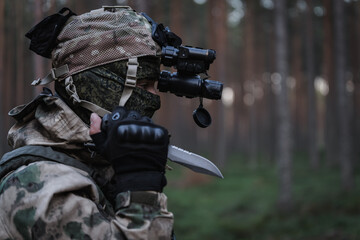 A soldier in balaclava camo, with a night vision device on his head and an knife in his hand in the...
