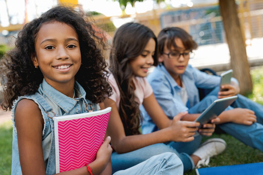 Portrait of preteen african school girl and children on background with backpacks and notebooks sitting at the park spend time outdoor backyard while break using gadgets tablet smart phone